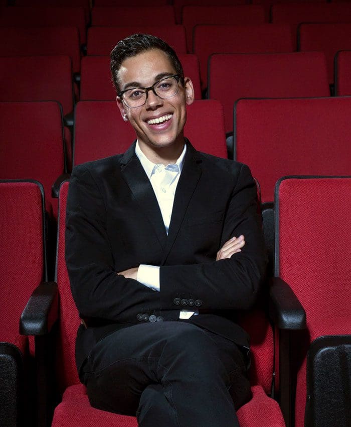 Evan Neff wearing black suit seated in movie theater with red seats